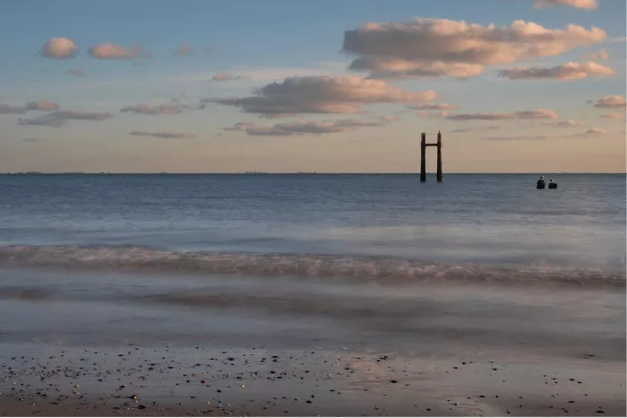 Kalme zee met een paal in het water en roze verlichte wolkjes in een blauwe lucht