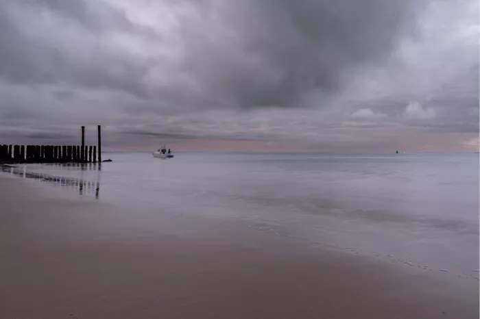 Stil strand met paalhoofden en een klein bootje op spiegelglad water onder grijze wolken