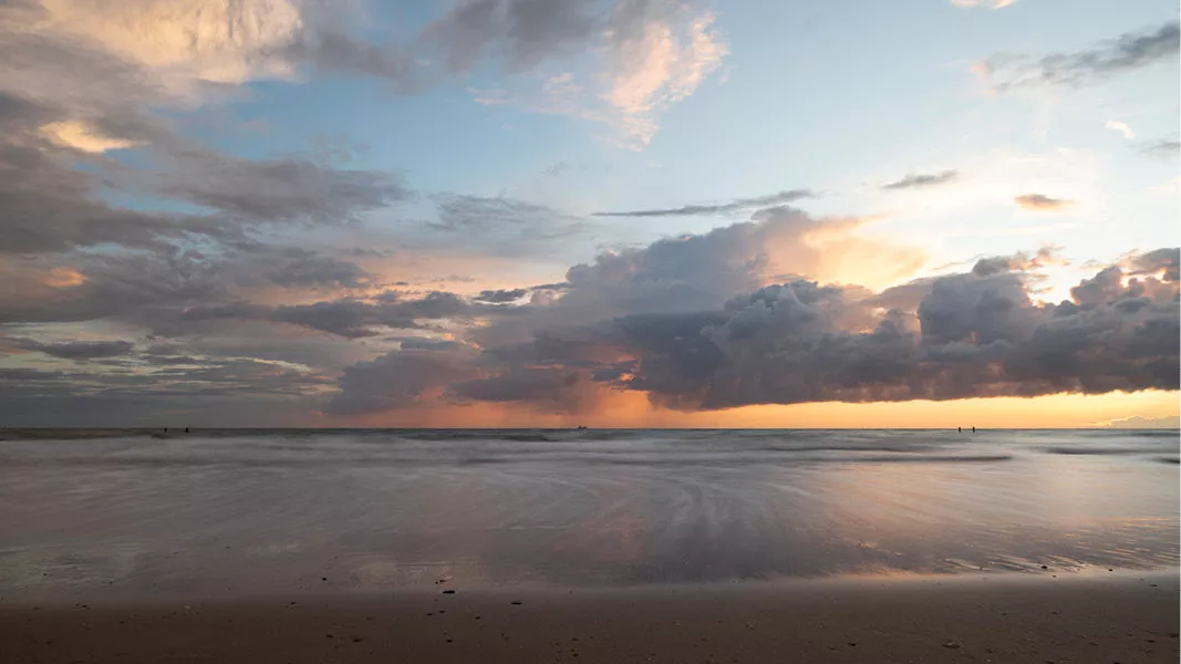 Zonsondergang aan zee met een grote wolkenbank, roze en gele kleuren en een nat glanzend strand