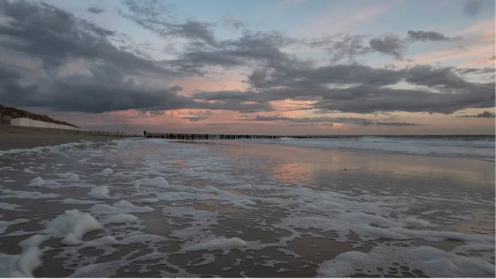 Verlaten strand bij zonsondergang met schuim op het zand, paalhoofden en strandhuisjes in de verte