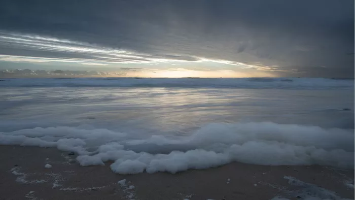 Zonsondergang onder donkere wolken met licht dat in de zee spiegelt en wit schuim op het strand