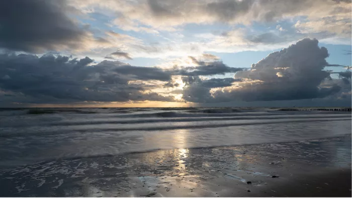 Zonsondergang tussen donkere stapelwolken met een glanzend lichtspoor over het natte strand