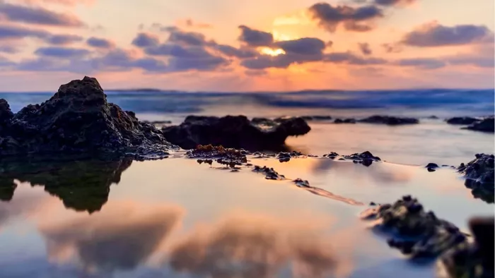 Gestileerd beeld van een zonsondergang aan de kust, met de zon die ondergaat aan de horizon. De lucht is gevuld met oranje, roze en paarse tinten, die worden weerspiegeld in het water op de voorgrond. Rotsformaties zijn zichtbaar langs de kustlijn, met de golven die zachtjes over de rotsen slaan.