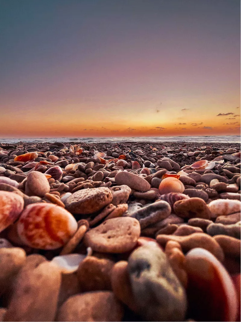 Close-up van een strand bezaaid met kiezels en schelpen bij zonsondergang. De zon is al onder, maar de hemel is nog steeds verlicht met een zachte oranje en paarse gloed.