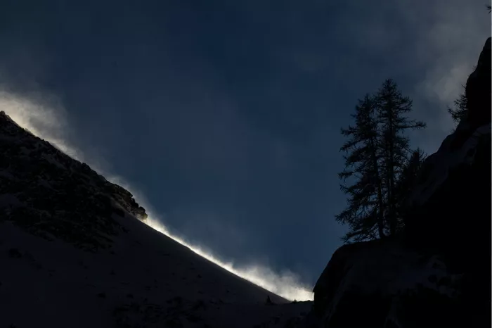 Opwaaiende sneeuw licht op in de avondzon langs een donkere berghelling, met naaldbomen als silhouet
