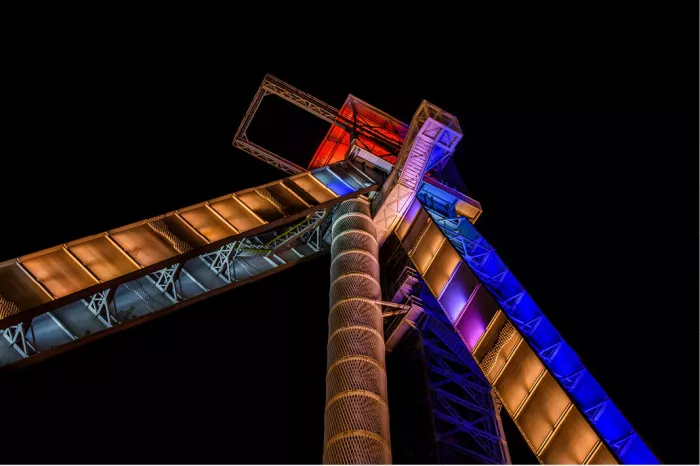 De iconische schachtbokken van C-Mine in Genk worden 's avonds sfeervol verlicht en steken krachtig af tegen de donkere hemel. Vanuit een laag standpunt benadrukt de compositie de indrukwekkende hoogte en industriële architectuur van dit voormalige mijncomplex.