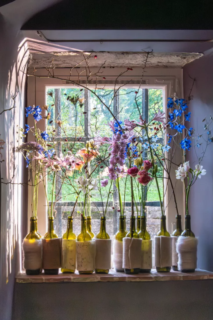 Wijnflessen met een wollen of vilten omwikkeling staan op een stenen vensterbank, elk met losse stelen van blauwe ridderspoor, roze nerine, witte alstroemeria en rozerode bloemen, met kale takken met rode besjes ertussen, terwijl zonlicht binnenvalt en het raam uitkijkt op een groene tuin.