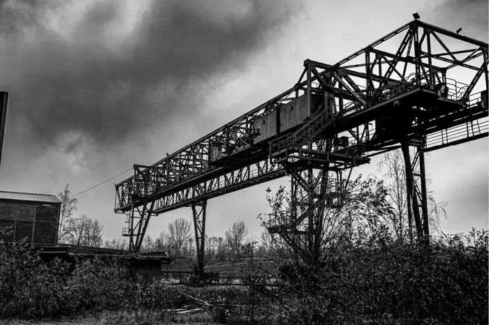 Grote metalen portaalkraan boven een verlaten, begroeid terrein onder een donkere wolkenlucht, in zwart-wit