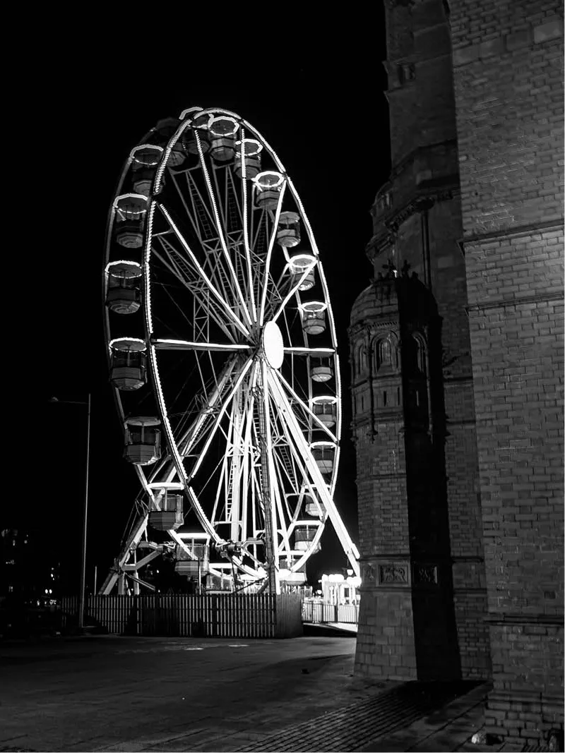 Verlicht reuzenrad bij nacht naast een bakstenen gebouw met ronde toren, in zwart-wit