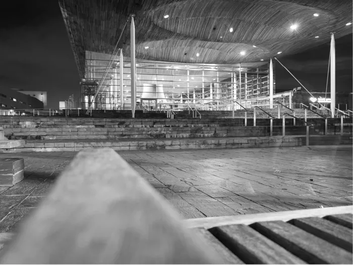 Modern gebouw met glazen gevel en golvend dak bij nacht, met trappen en een houten bank vooraan, in zwart-wit