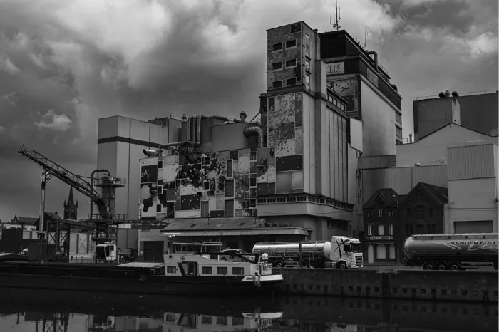 Fabriek met silo's en kraan aan een kanaal met een binnenschip en tankwagens, in zwart-wit