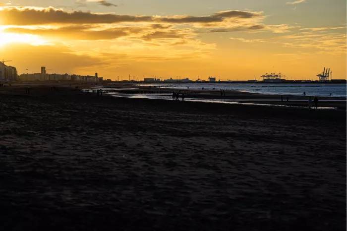 Strand bij zonsondergang met gouden wolken, gebouwen links en havenkranen aan de horizon