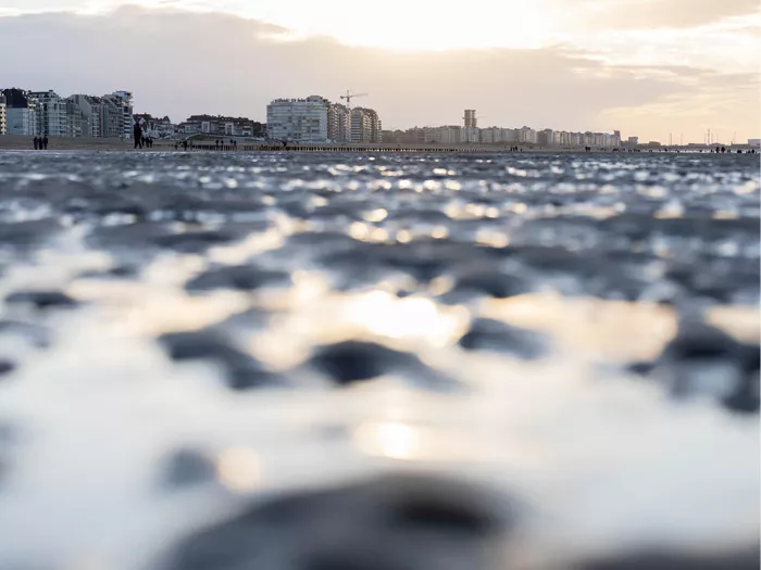 Nat zand met ribbels en lichtreflecties van dichtbij, met gebouwen aan de horizon