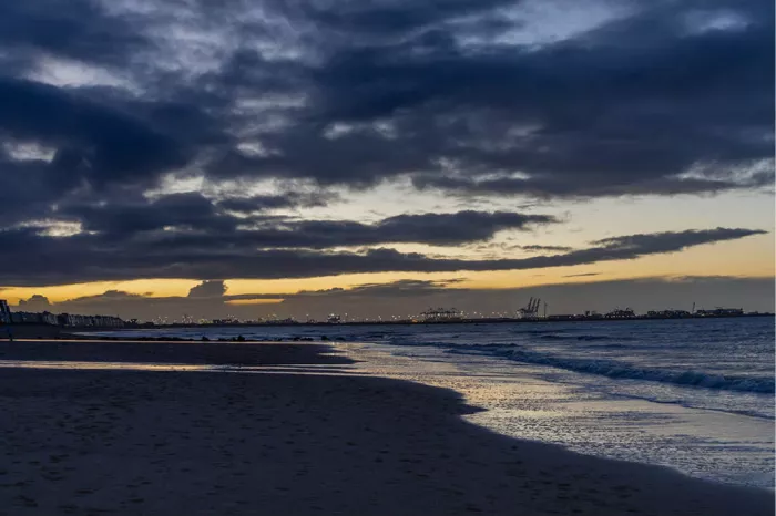 Strand bij schemering met donkere wolken, een gele lichtband en golven op het natte zand
