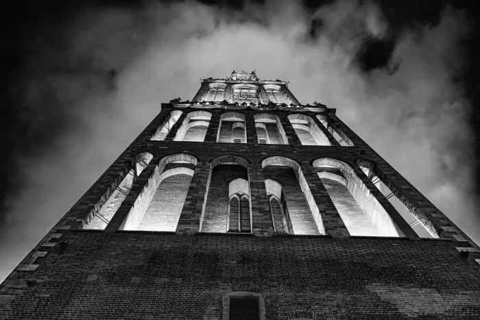 Laag standpunt langs een verlichte bakstenen toren met hoge spitsboogvensters die bij nacht oprijst tegen een zwaar bewolkte lucht, in zwart-wit.