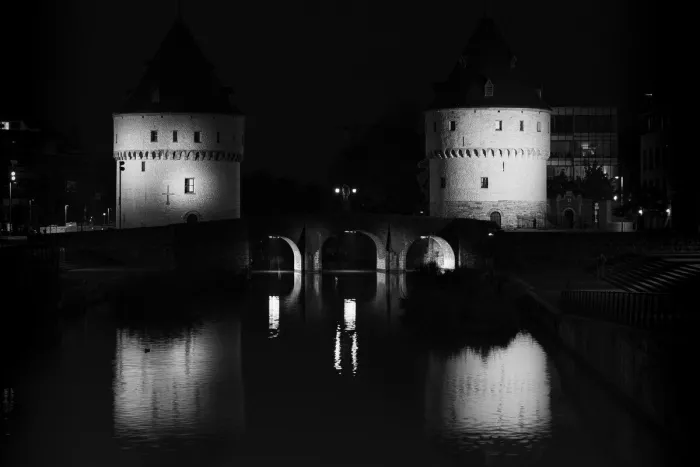 Twee verlichte ronde stenen torens met kegelvormige daken staan weerspiegeld in het donkere water van een rivier, met een boogbrug ertussen en een zwarte nachthemel erboven.