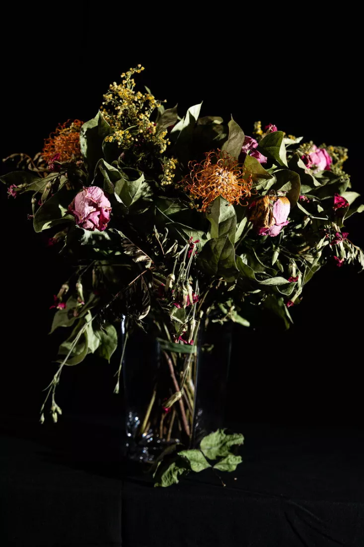 Verwelkt boeket met gedroogde roze rozen, oranje naaldkussenprotea, gele guldenroede en groene bladeren in een glazen vaas, fel belicht tegen een zwarte achtergrond en op een zwarte doek.